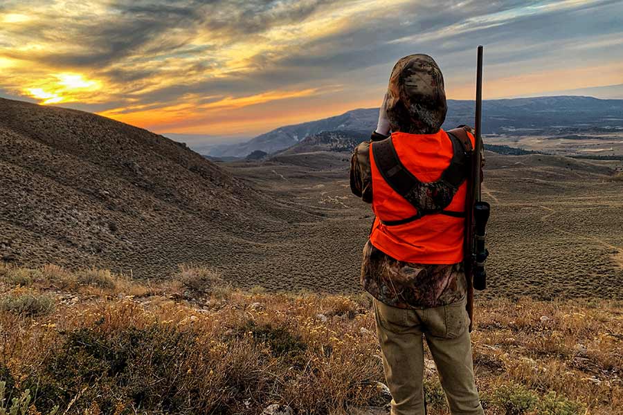 Hunter carrying a rifle and scope looking over a wide valley at sunset