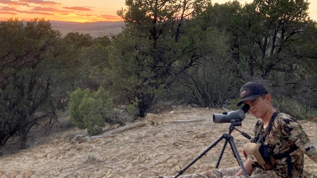 A youth hunter sitting on the ground, peering at wildlife through a scope