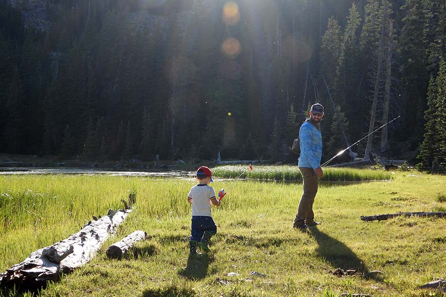 A father and his young son walking through the forest to a fishing pond, carrying poles and gear